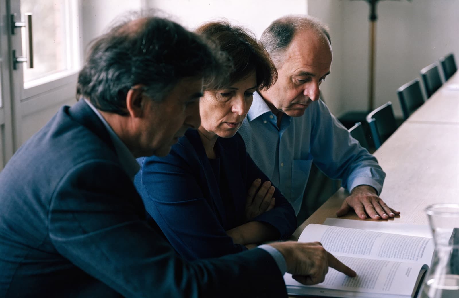 Trois dirigeants penchés sur un rapport imprimé ouvert sur la table d'une salle de réunion, l'un pointant un paragraphe du doigt.